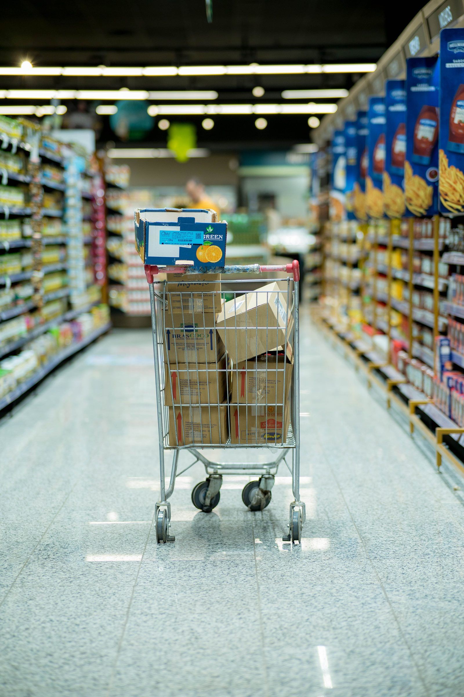 Un carrello carico di scatole fermo in una corsia di supermercato, visto da dietro, tra scaffali sfocati.